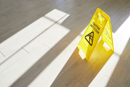 Yellow caution sign reading “Wet Floor” placed on a bright wooden floor with sunlight casting shadows.